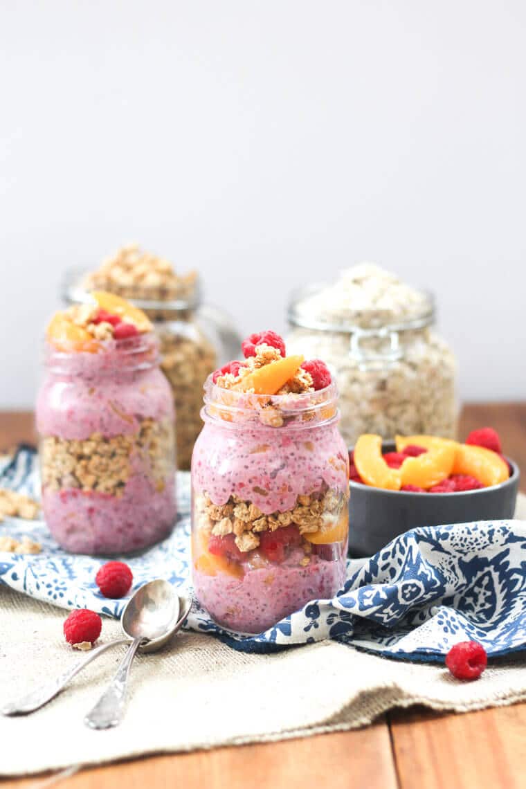 Two mason jars containing peach melba crumble overnight oats with a jar of oats and bowl of peaches in the background.