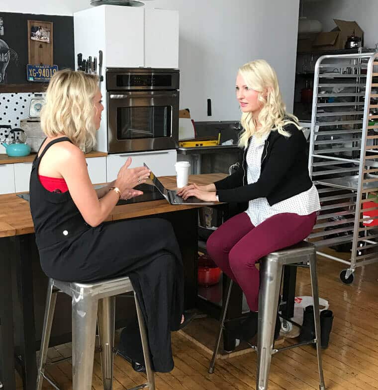 Abbey Sharp sitting at a bar stool typing on a laptop interviewing a woman.