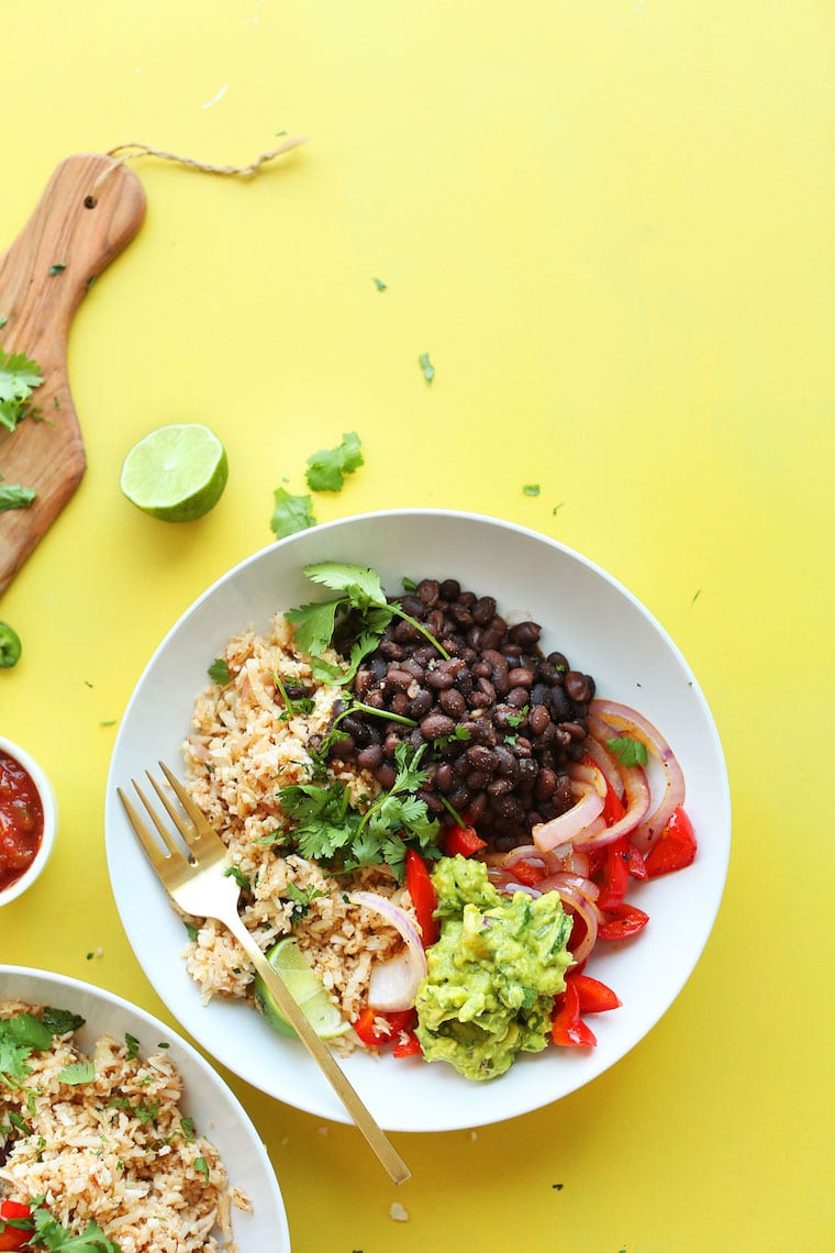 A white plate filled with beans, rice, veggies and guacamole on a bright yellow surface.