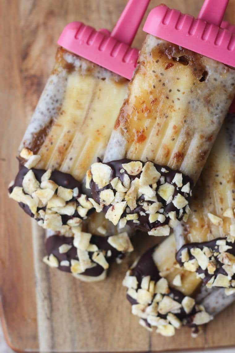 Banoffee pie popsicles coated in chocolate and banana chips on a wooden platter.