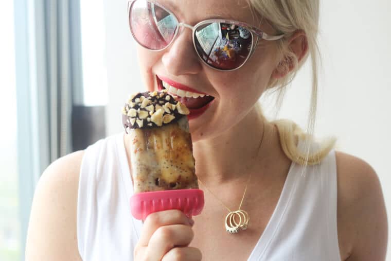 Women biting into a banoffee pie popsicle coated with chocolate and banana chips.