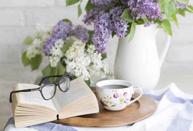 A bouquet of flowers in a vase on a table with a book, glasses, and mug on top.