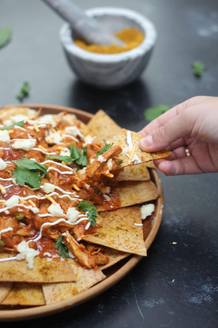 A hand lifting up a chip from a plate of butter chicken nachos.