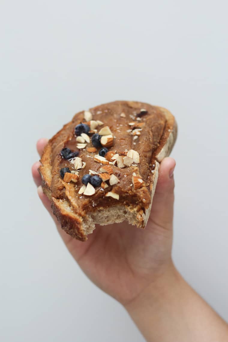 A piece of bread with salted caramel almond butter spread on top with blueberries on top being held up by a hand.