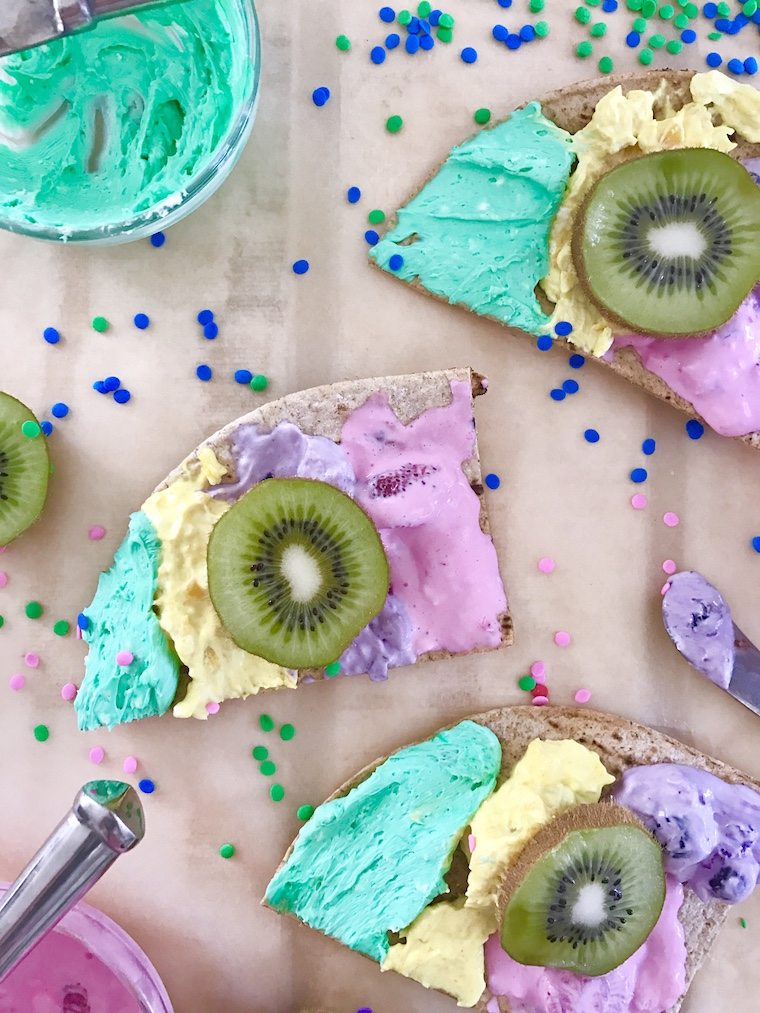 A piece of toast with teal, yellow, and purple spread with a kiwi on top.