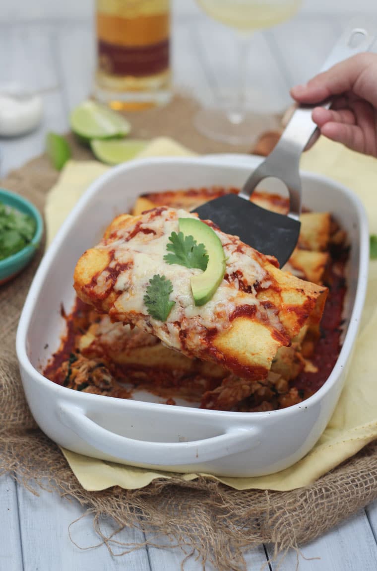 A white baking dish with chicken and butternut squash enchiladas with a spatula lifting a serving up.