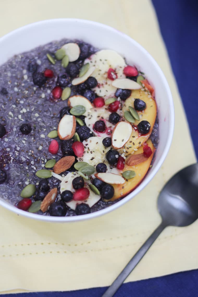 An overhead image of a white bowl of blueberry quinoa with fresh fruit with nuts and seeds on top.