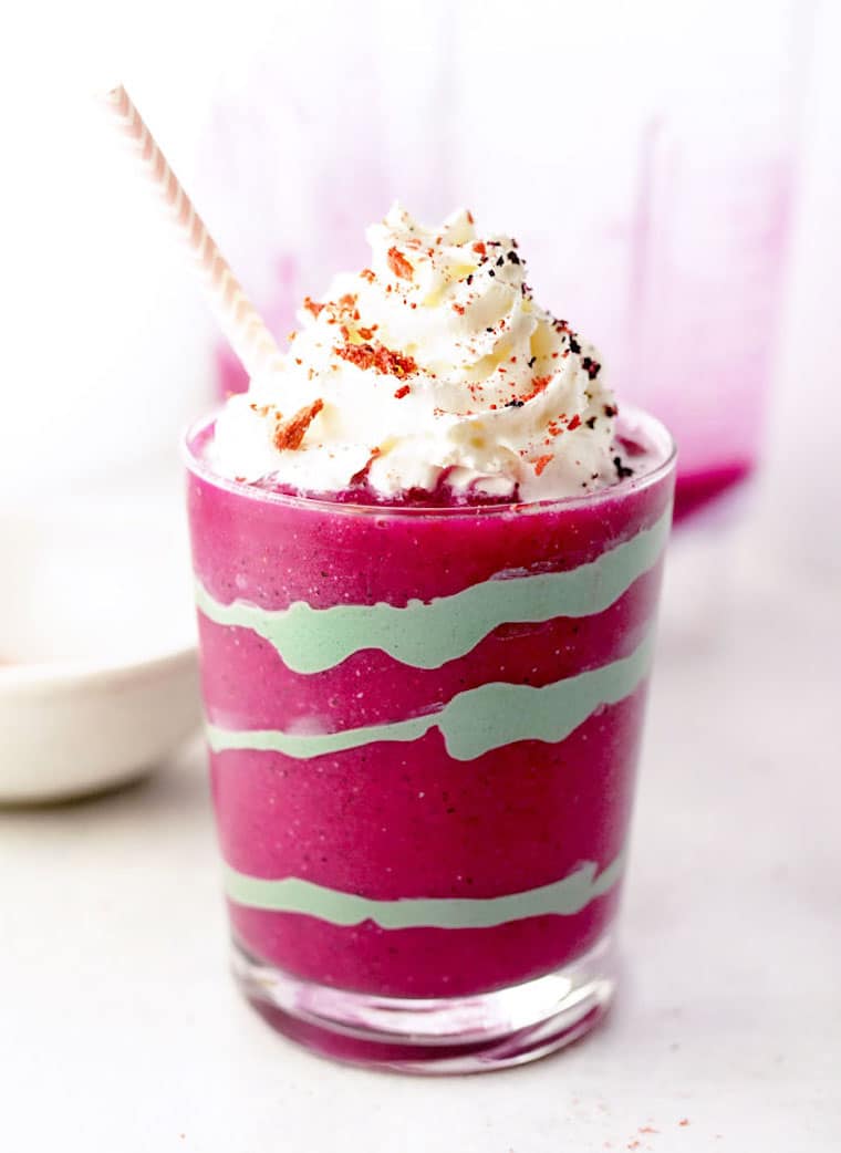 A glass of raspberry coloured and light green swirled smoothie with whipped cream on top.