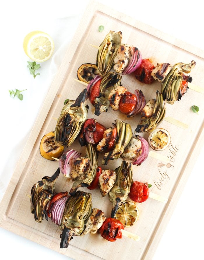 birds eye view of mediterranean chicken and artichoke kebobs garnished with lemon on a wooden serving board 