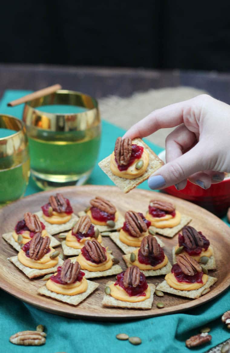 A plate of sweet potato and cranberry mousse bites with a hand lifting up a single one and two glasses in the background.