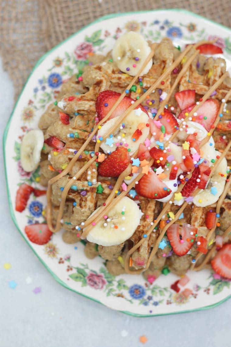 A close up overhead view of a stack of birthday cake protein waffles on a white plate with strawberries and sprinkles on top.