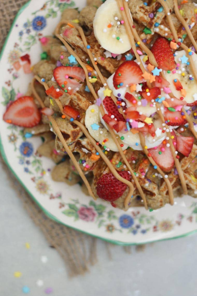 A close up overhead view of a stack of birthday cake protein waffles on a white plate with strawberries and sprinkles on top.