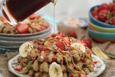 A stack of birthday cake protein waffles on a white plate with strawberries on top with a bottle of syrup being poured on.