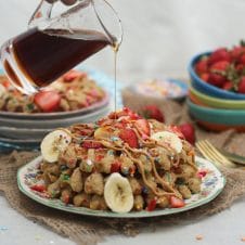 A stack of birthday cake protein waffles on a white plate with strawberries on top with a bottle of syrup being poured on.