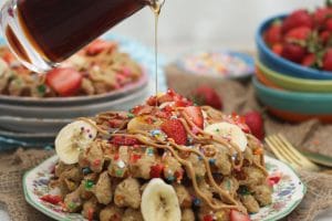 A stack of birthday cake protein waffles on a white plate with strawberries on top with a bottle of syrup being poured on.