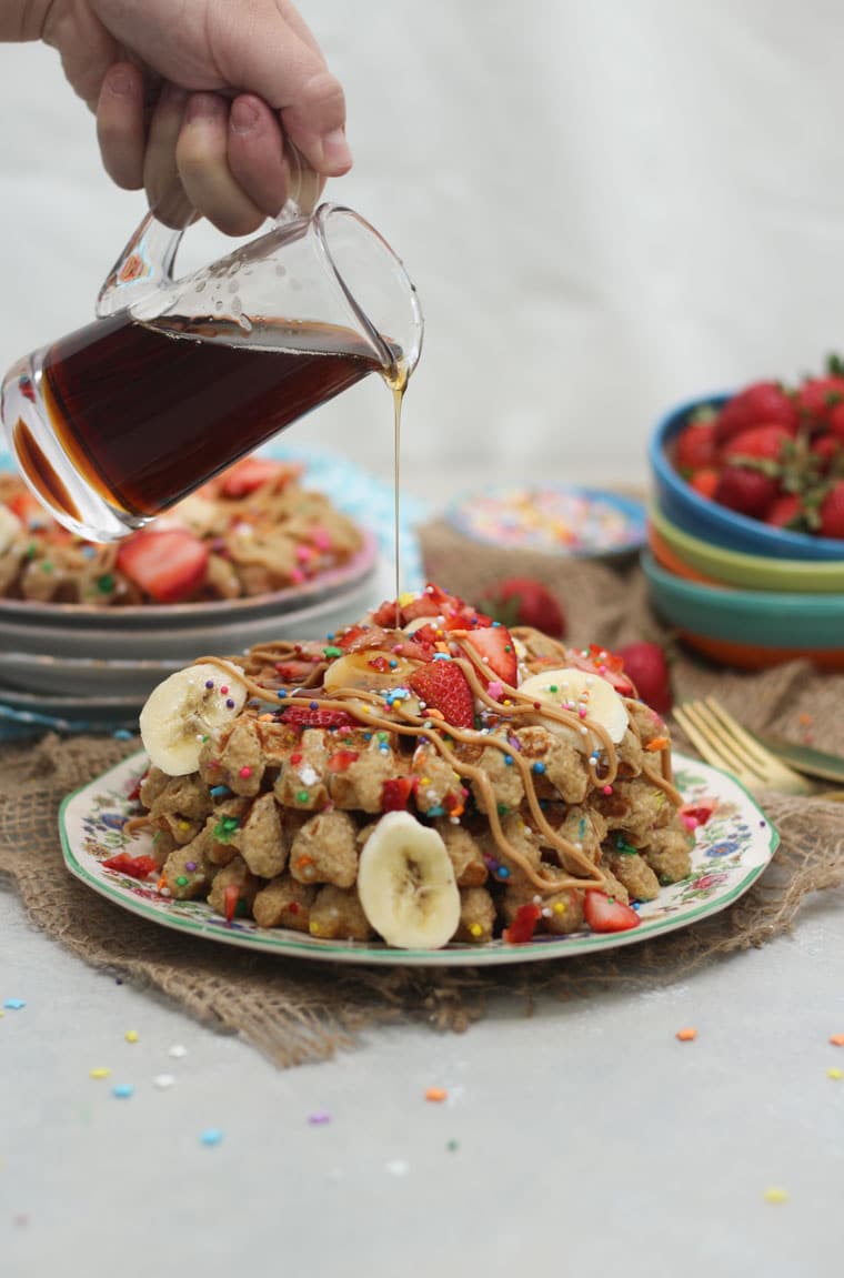 A stack of birthday cake protein waffles on a white plate with strawberries on top with a bottle of syrup being poured on.