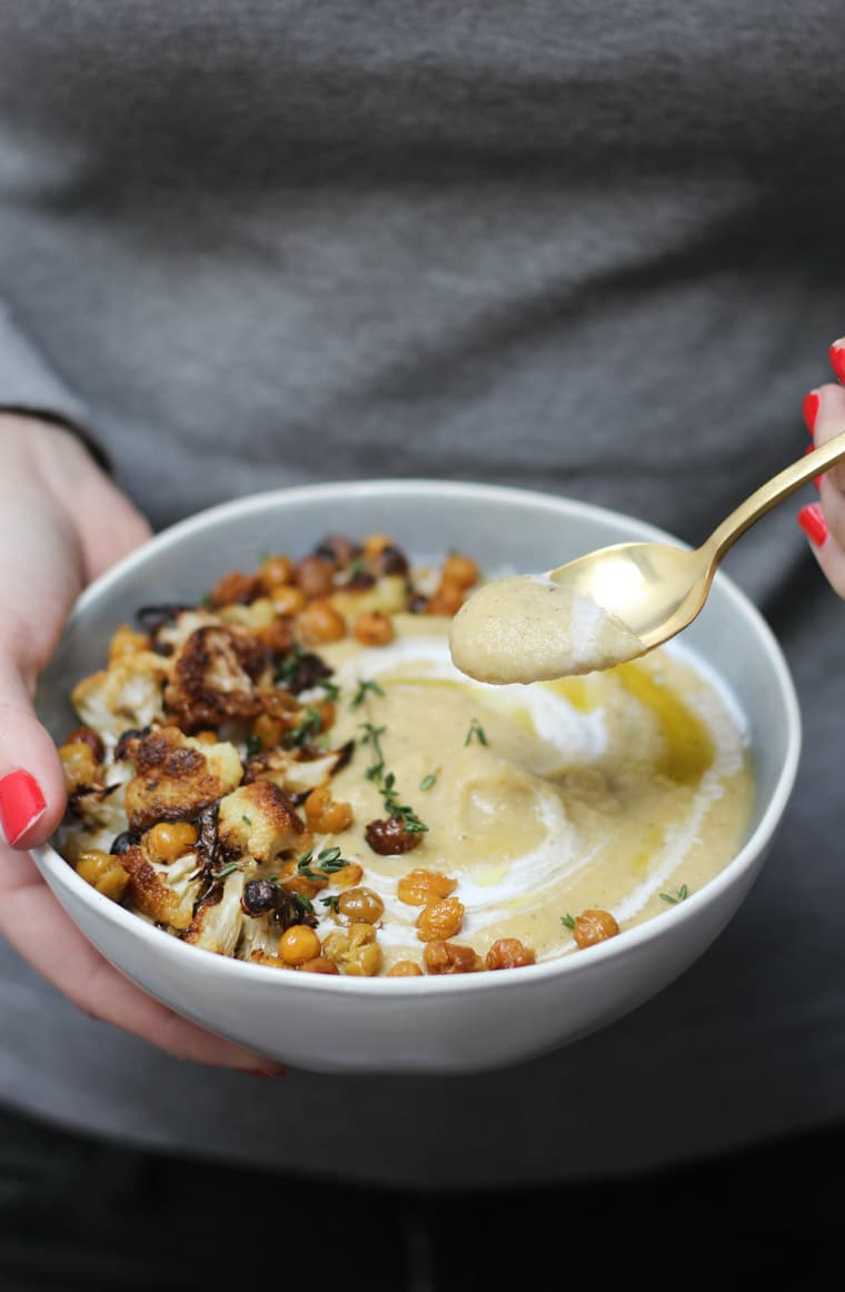 Hands holding bowl of vegan cauliflower and parsnip soup garnished with fresh herbs and chickpeas and spoon.