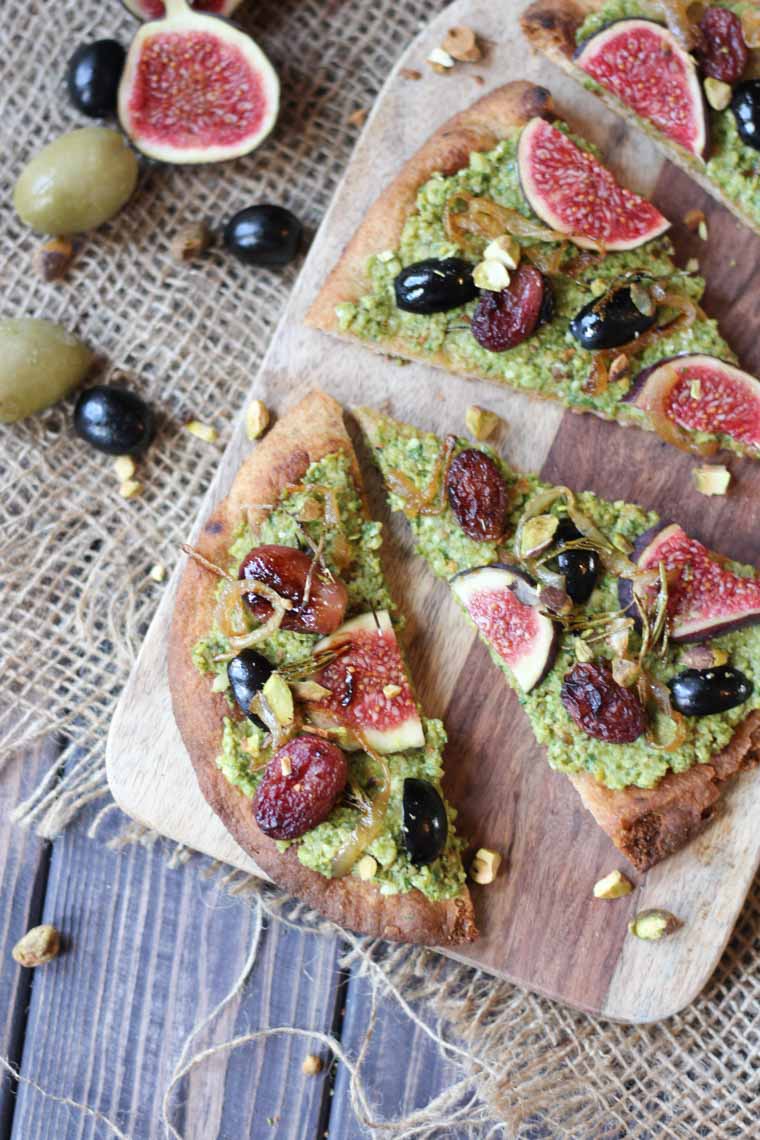 birds eye view of vegan pesto and olive flatbread on a wooden serving board topped with figs and roasted grapes