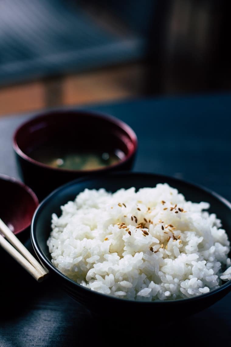 A bowl of rice in a dark bowl with miso soup in the background.