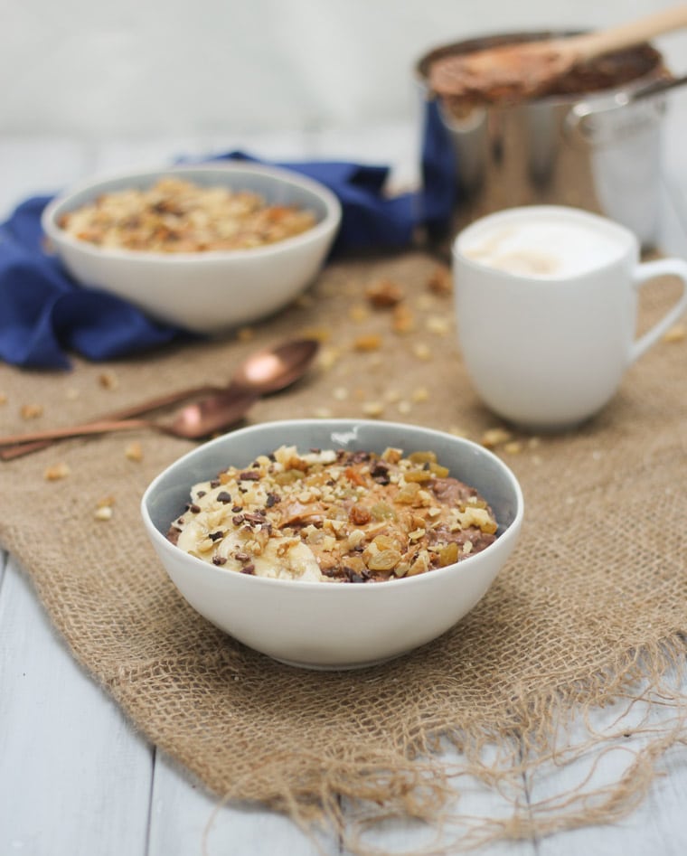 An angled photo of a bowl of chocolate zucchini bread oatmeal zoats with bananas and chocolate chips on top on a dining room table with a mug of coffee in the background.