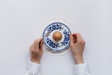 An overhead photo of an egg on a plate with hands around it, about to dig in with a spoon.