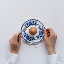 An overhead photo of an egg on a plate with hands around it, about to dig in with a spoon.