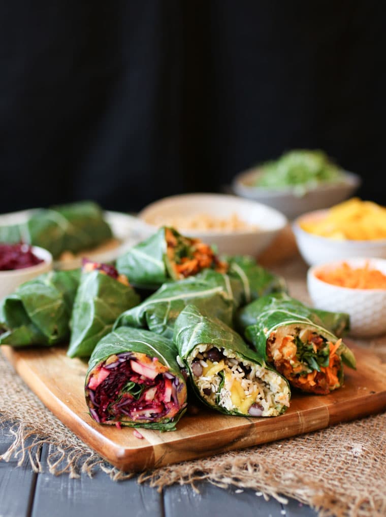 three vegan collard green wraps with assorted fillings served on a wooden platter with additional collard wraps and bowls of vegetables in the background