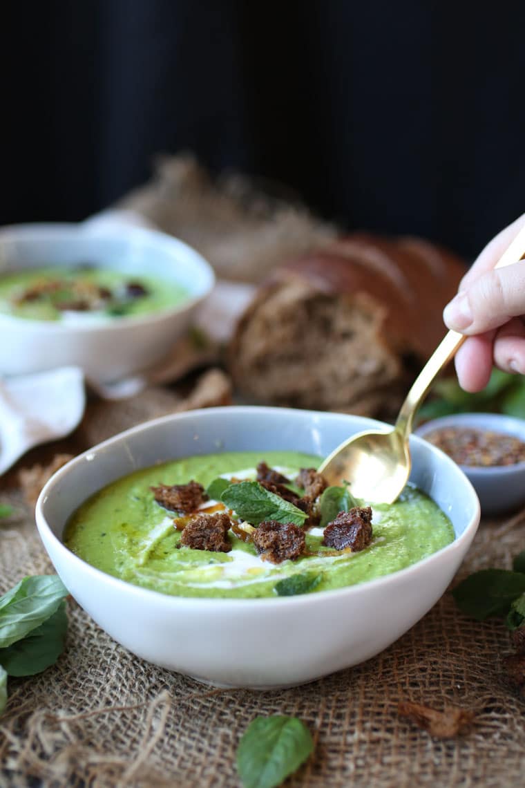 A white bowl with pea, mint, and coconut soup with a gold coloured spoon scooping into it.