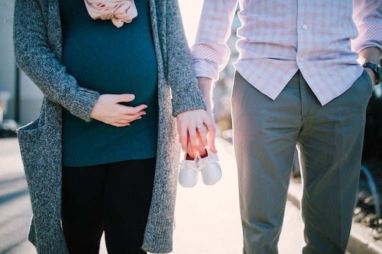 A pregnant woman holding the hand of her partner and a pair of baby shoes in between, discussing what not to say to a pregnant woman about her body.