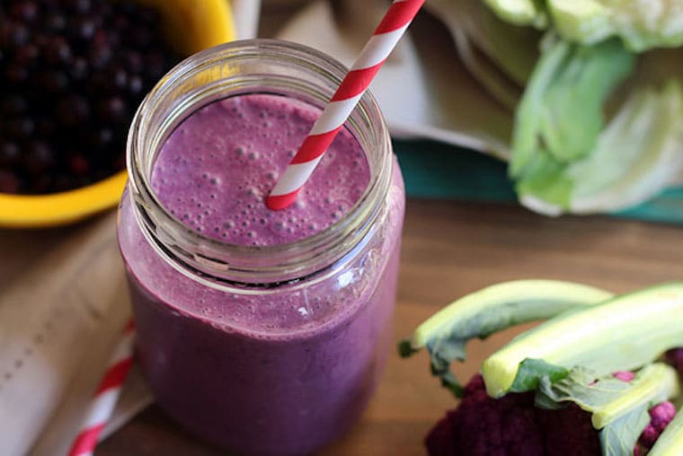 close up of vegan and gluten free wild blueberry and cauliflower smoothie served in a clear jar with a red and white straw with fresh fruit and cauliflower in the background