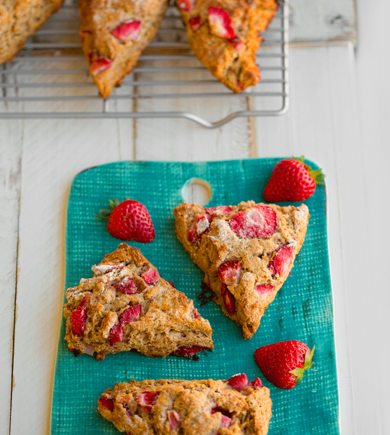 three vegan strawberry sunrise scones for easter brunch served on a blue plate garnished with fresh strawberries and additional strawberry scones on a cooling rack in the background