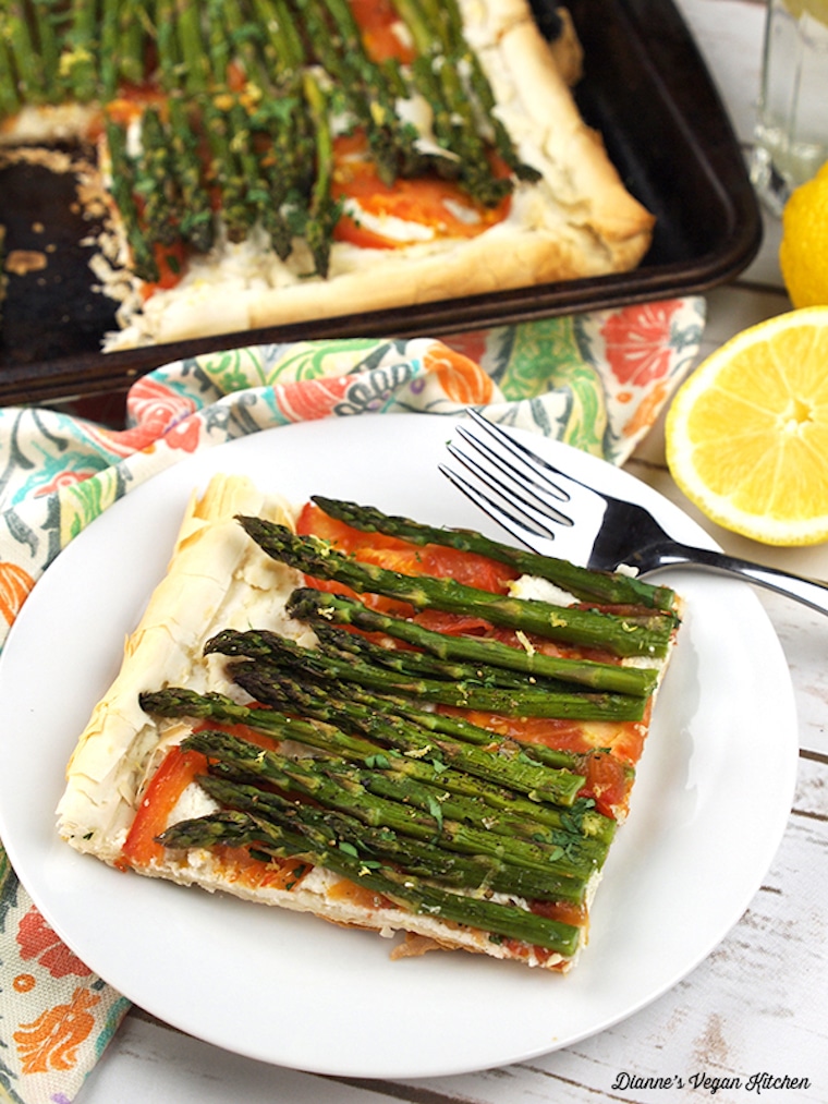 slice of vegan asparagus and tomato tart with cashew ricotta on a white plate with a silver spoon with the rest of the tart on a black baking tray in the background and fresh lemon wedges on the side