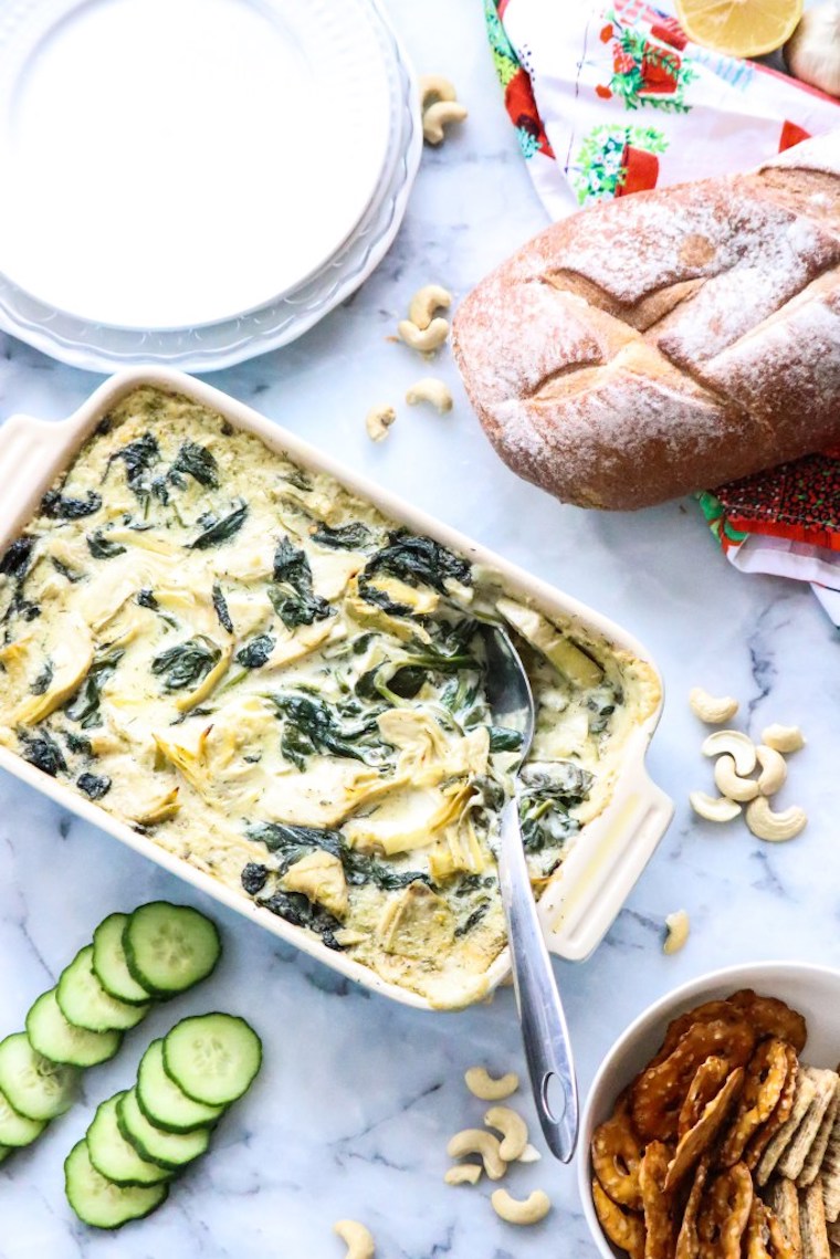 birds eye view of creamy vegan artichoke spinach dip in a white casserole dish next to a loaf of bread, cashews, pretzels, and fresh cucumbers on a marble counter top