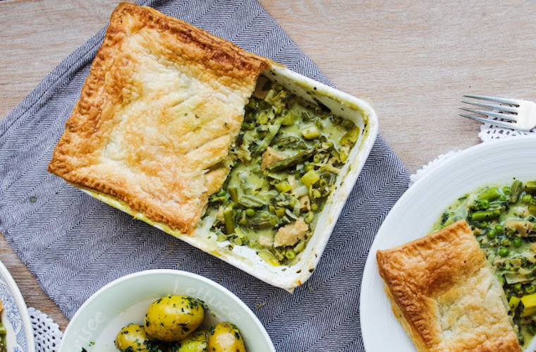 birds eye view of vegan wild garlic and spring vegetable pot pie in a white casserole dish next to a slice of savoury pie on a white plate and a white bowl containing herbed potatoes