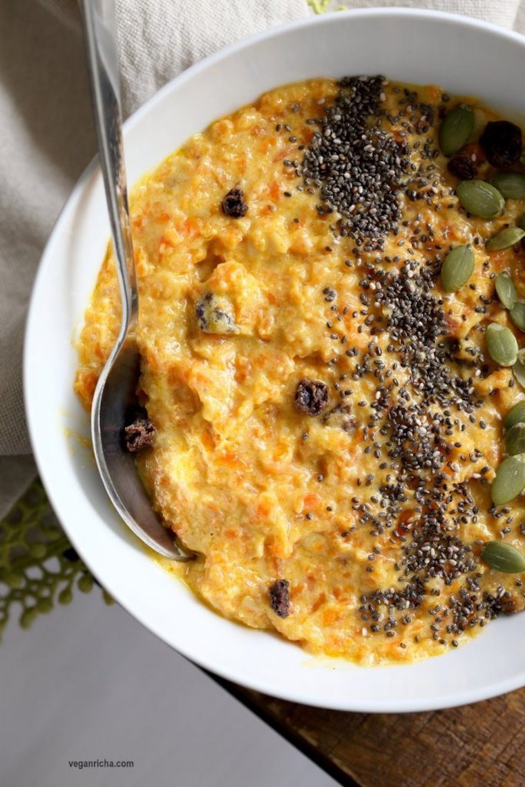 close up of vegan and gluten free carrot pudding with cardamom in a white bowl with a silver spoon garnished with various seeds