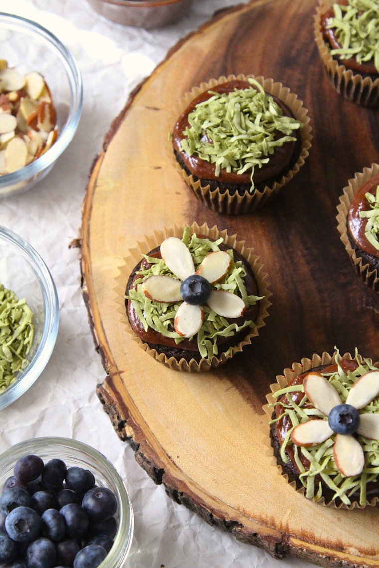 birds eye view of vegan chocolate easter cupcakes garnished with almonds and blueberries on a wooden serving board next to clear bowls with additional garnished inside