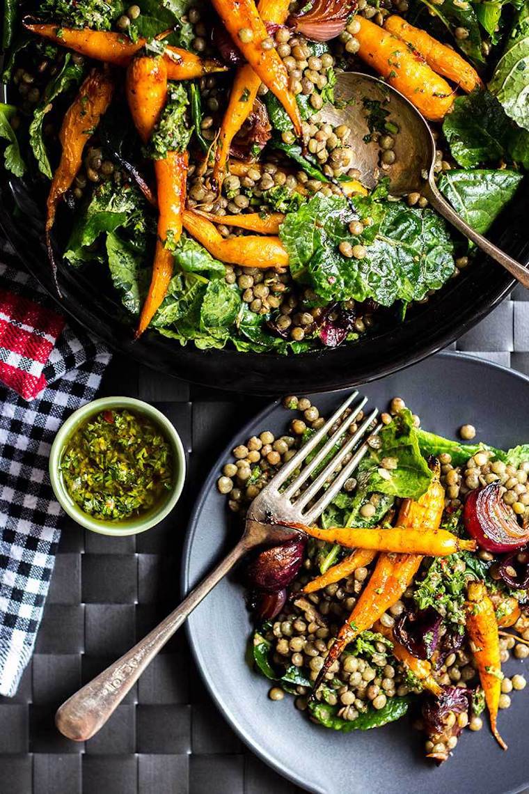 birds eye view of vegan and gluten free lentil salad with roasted baby carrots and red onions on a grey plate with a silver fork next to a black bowl containing additional lentil salad and a small bowl containing herb dressing
