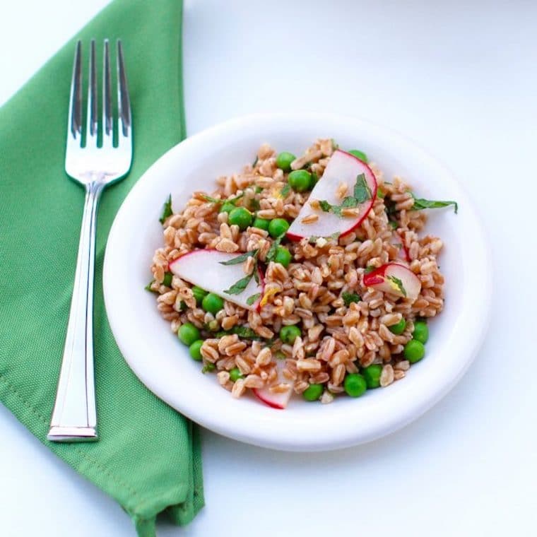 vegan spring pea and radish farro salad garnished with lemon and mint vinaigrette in a white bowl next to a green napkin with a silver fork on top