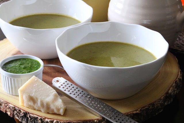 image of two white bowls containing green vegan asparagus spinach soup with parmesan cheese and green pesto on the side