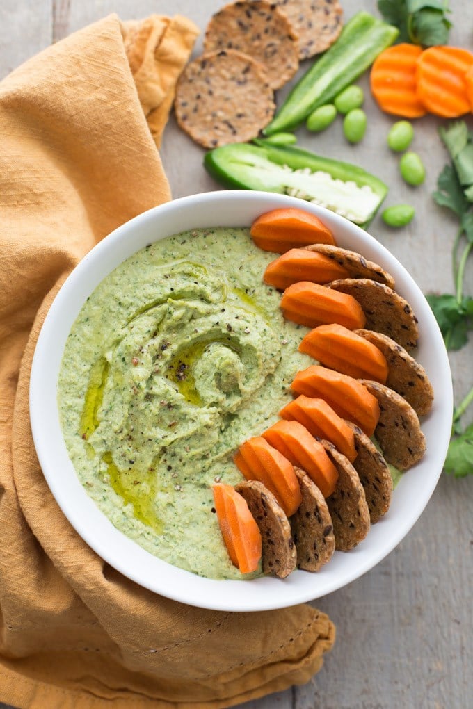 birds eye view of vegan green cilantro jalapeno edamame hummus garnished with crackers and carrot slices in a white bowl