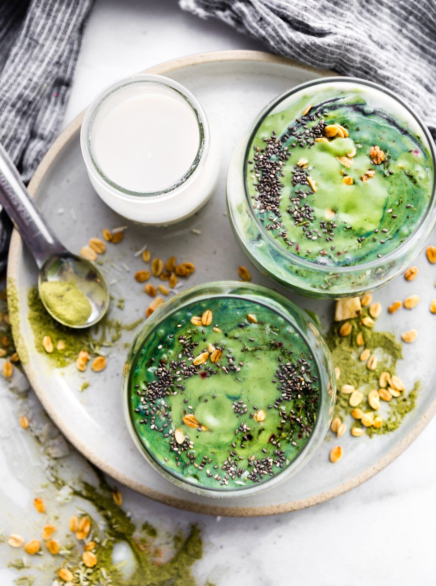birds eye view of two bowls filled with vegan coconut spirulina green smoothies topped with granola and seeds and a clear glass of coconut milk on the side 