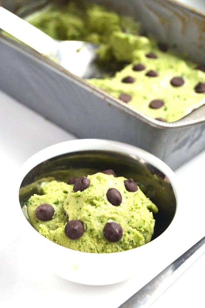 close up of vegan mint chocolate chip protein ice cream garnished with additional chocolate chips served in a white bowl with additional ice cream in a grey tray in the background