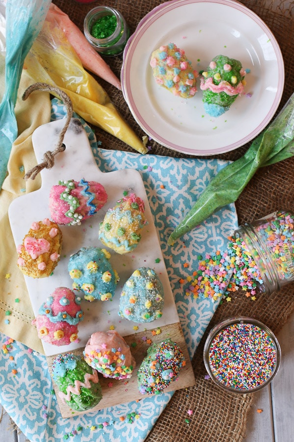 Overhead view of eight Vegan Gluten Free Rice Krispies Easter Eggs coated in a Homemade Vegan White Chocolate with sprinkles and candies on top on a serving board on top of a festive tablecloth