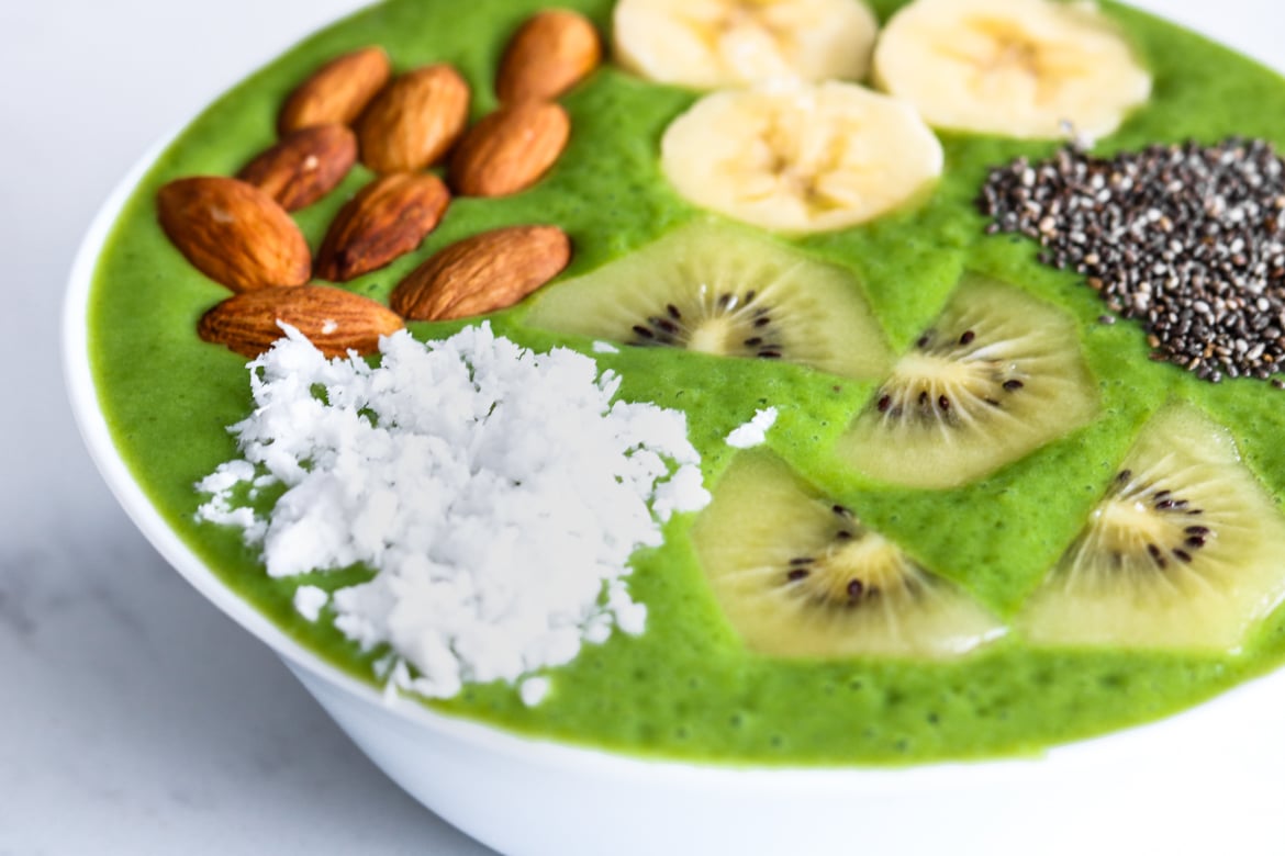 close up of vegan superfood green smoothie bowl topped with coconut, fruit, nuts, and seeds in a white bowl for st. patrick's day