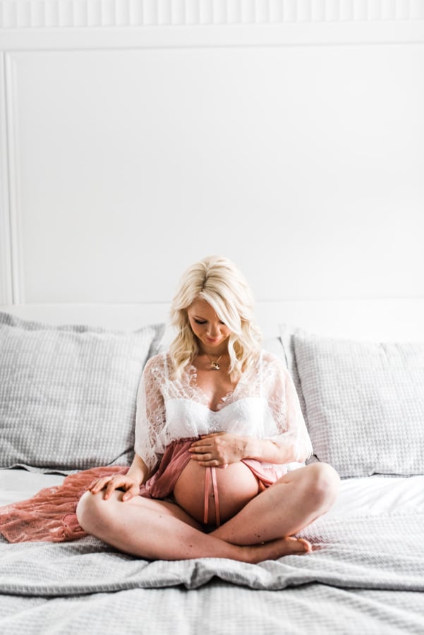 Pregnant Abbey sitting on a bed cross legged while holding her stomach.