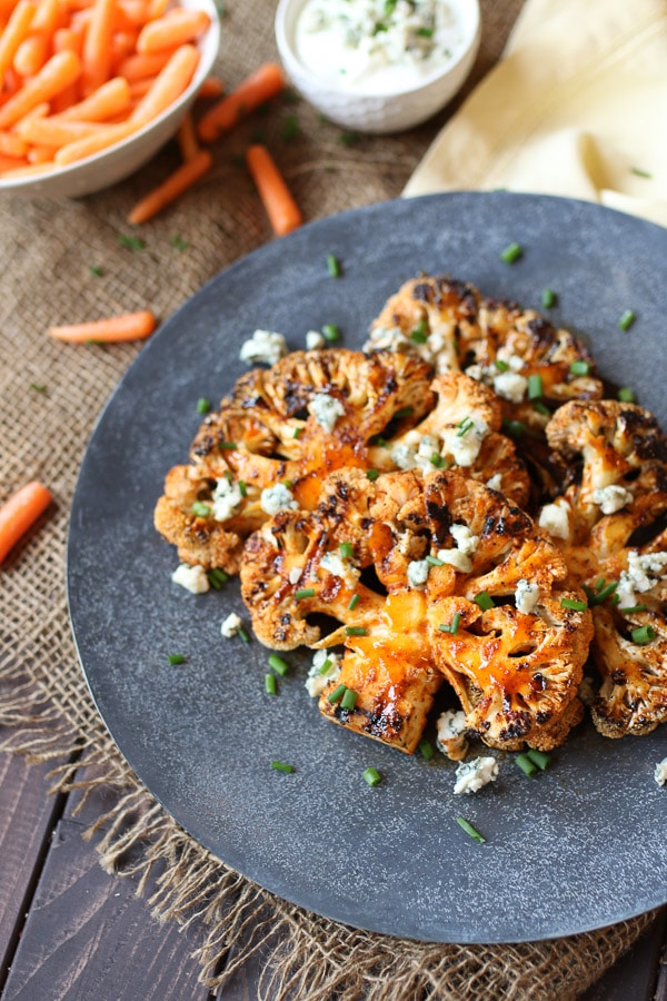 Buffalo cauliflower steaks on a grey serving plate topped with blue cheese and chives next to a bowl of carrots.