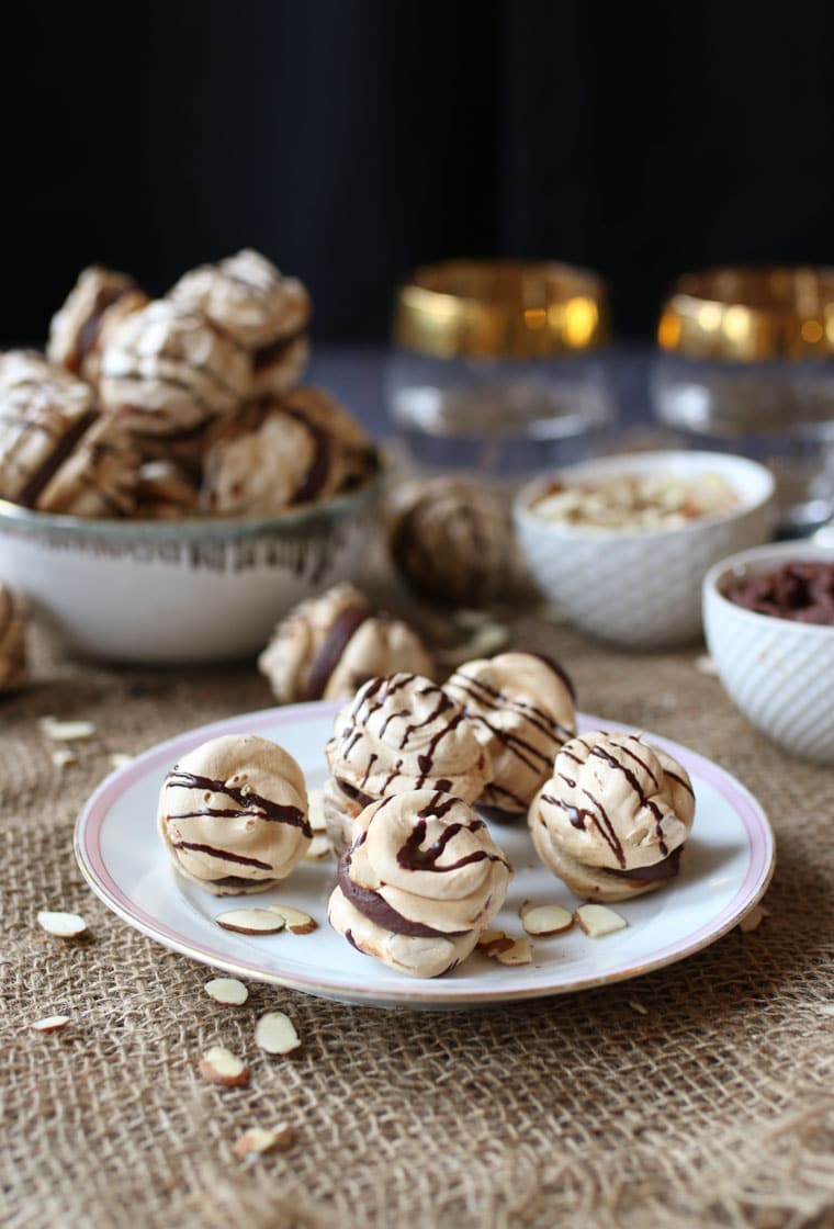 A plate with almond cherry chocolate meringue cookies with a chocolate ganache with a bowl of them in the background.