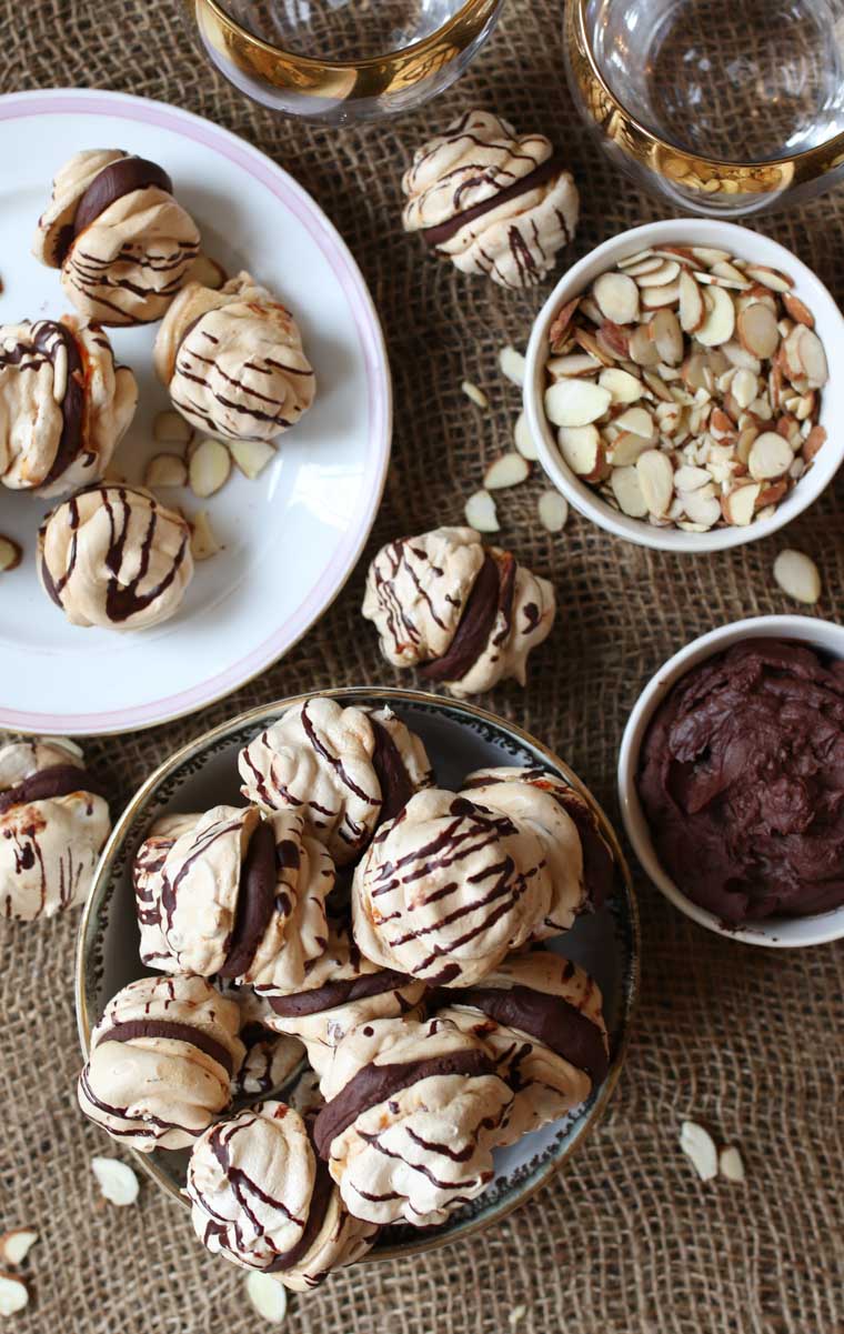 Two plates of almond cherry chocolate meringue cookies with a chocolate ganache with a bowl of almond silvers, and bowl of chocolate.