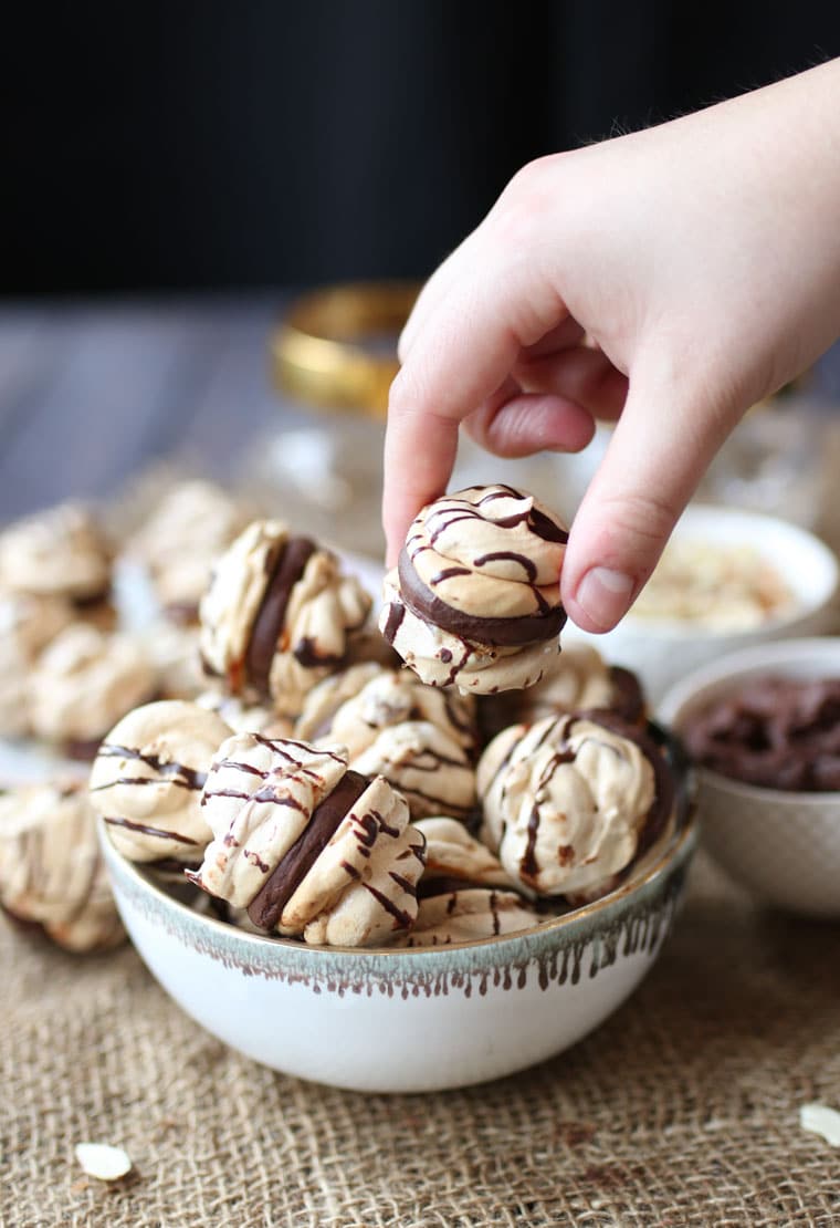 A hand lifting up an almond cherry chocolate meringue cookies with a chocolate ganache from a bowl of them.