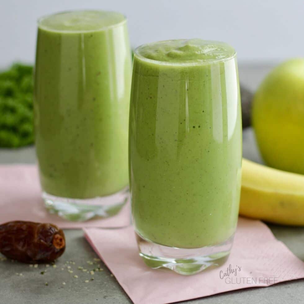 two tall clear glasses filled with vegan and gluten free green smoothies on pink napkins with fresh fruit in the background